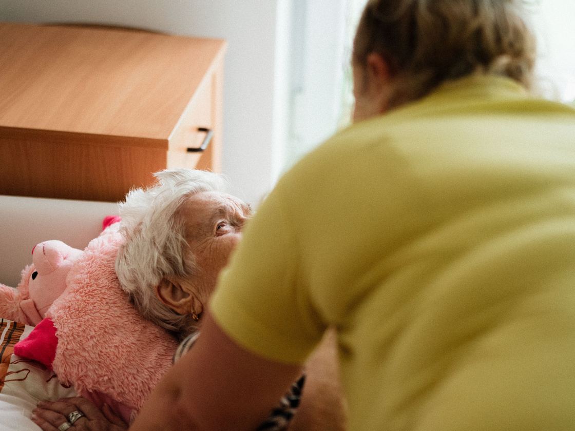 A caregiver helping an older woman sit up in bed at home.