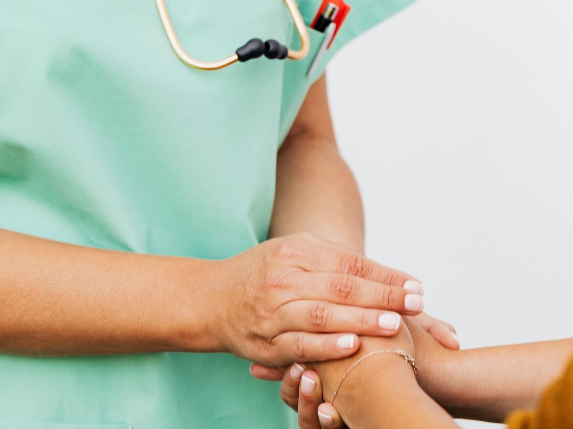 A nurse in scrubs holding an older person’s hand in both of hers.