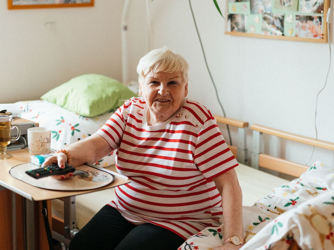 An older woman sitting on the edge of her bed at home, with her own photographs on the wall behind her.